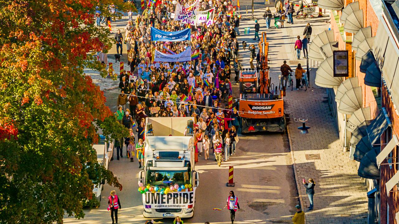 Drönarbild av en parad av människor som går i Pride-tåget i Umeå 2024, med regnbågsflaggor och banderoller.