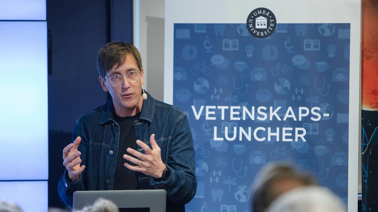 Föreläsare pratar inför publik, rollup med ordet vetenskapsluncher syns i bakgrunden.