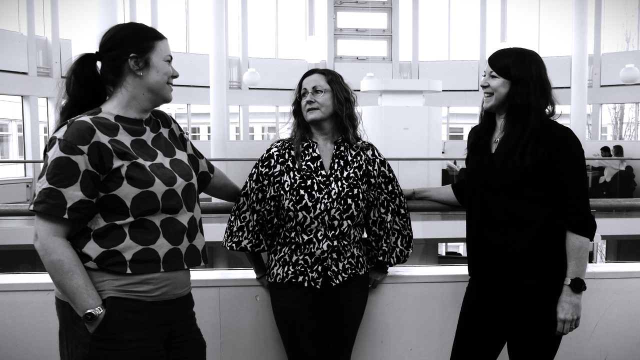 Tre kvinnor står och pratar och skrattar i en inomhusmiljö. Kortet är svartvitt.