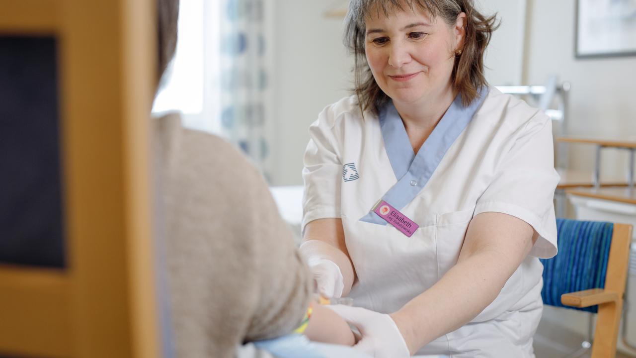 En person sitter med ryggen mot kameran och håller fram armen. Mitt emot sitter studiekoordinatorn Elisabeth Müller Granberg i färd med att ta ett blodprov.