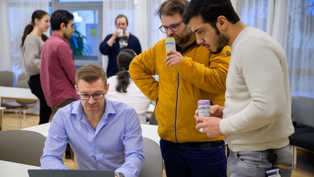 Anders Garpebring sits at a white table and looks at a laptop while two men hold beverages and look down at the screen.