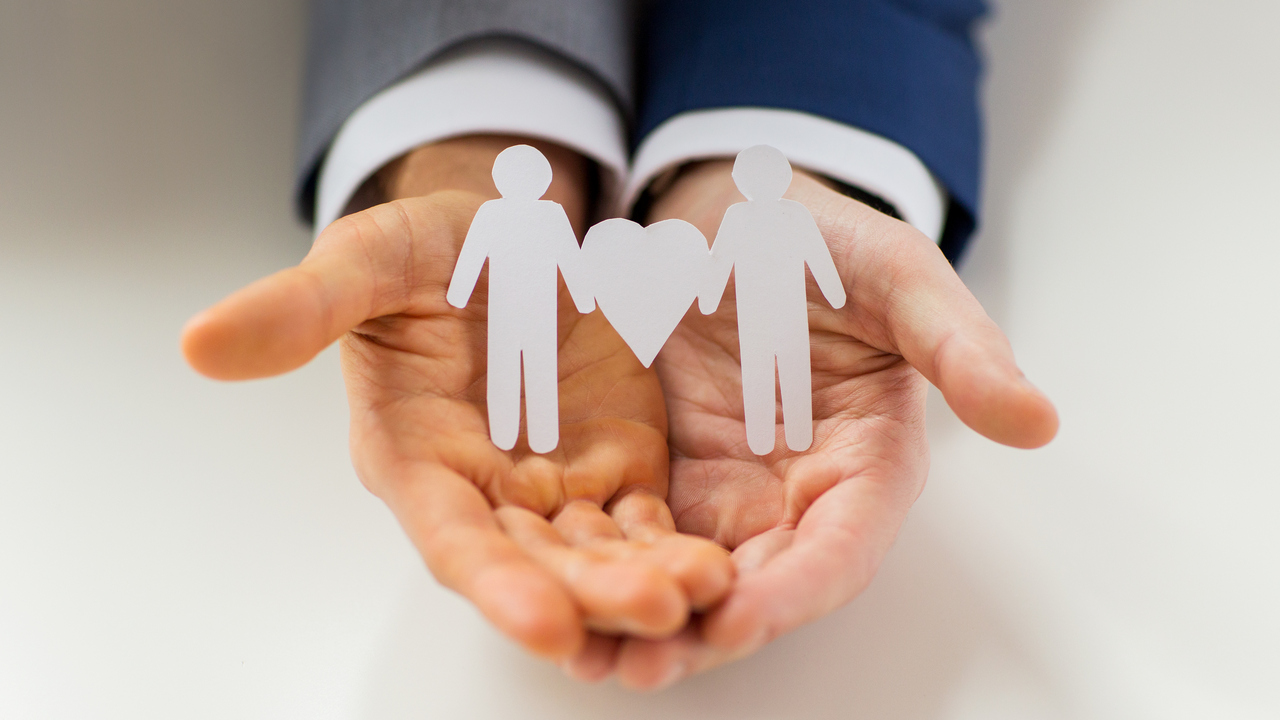 teHänder som håller två kvinnofigurer i papper med ett hjärta mellan. För att illustrera samkönad kärlek.st