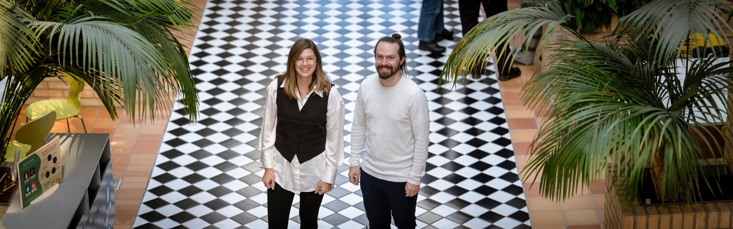 Porträtt på två forskare som står framför ett rutigt golv och mellan växter