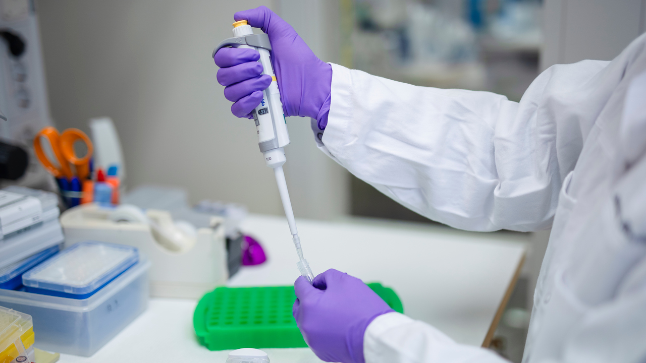 Närbild av armar som håller en pipett i den ena handen och ett mikrocentrifugrör i det andra. Spetsen har just stuckits ned i röret för pipettering.