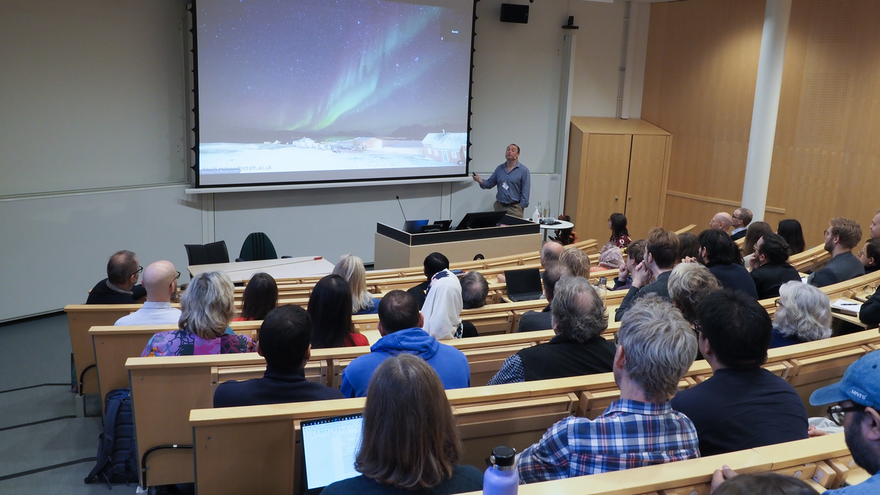 Hörsal fylld med människor som sittandes kollar mot talaren. Talaren visar upp en bild på norrsken på en stor projektorskärm.