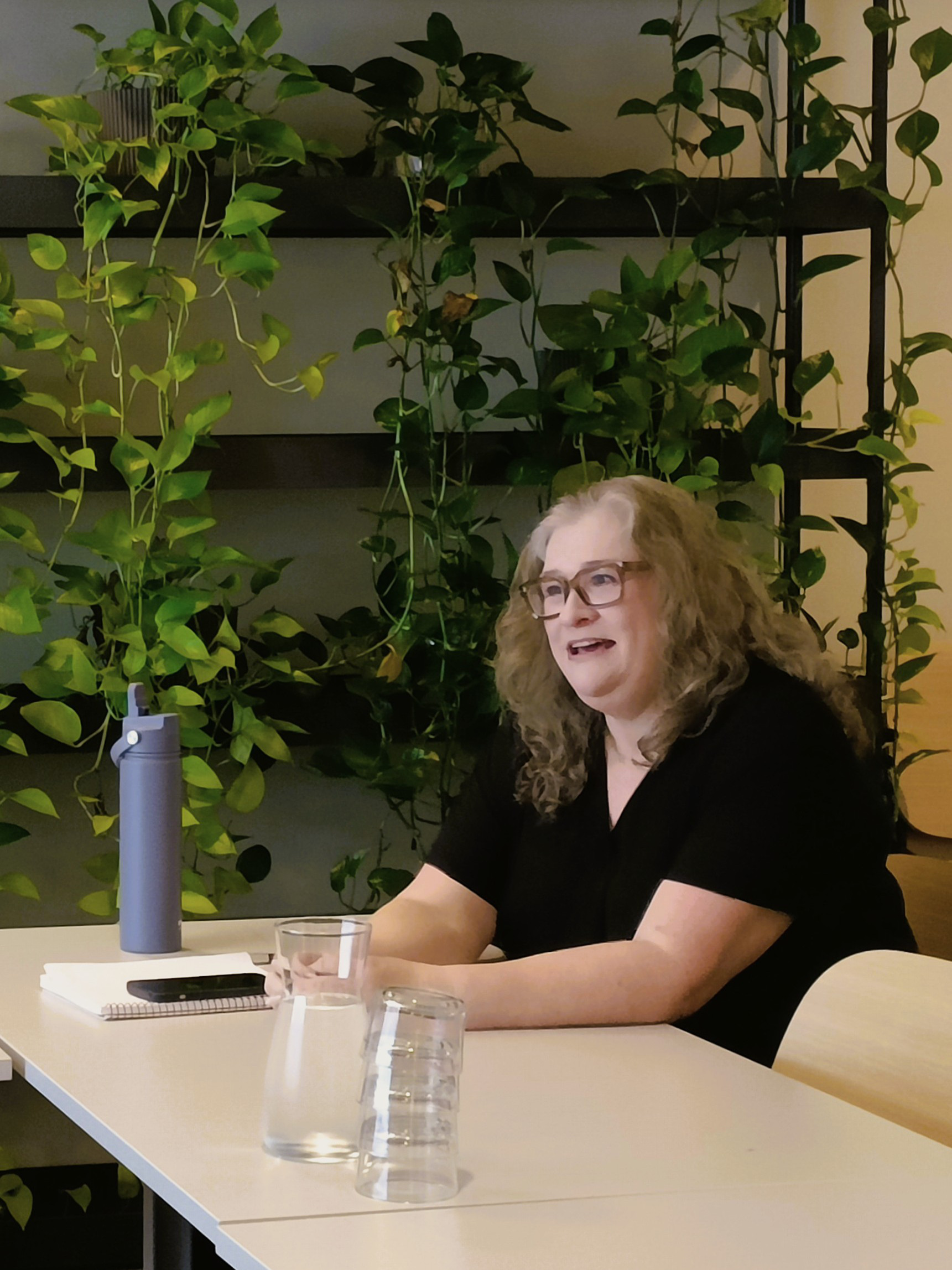Paula Wood-Adams sitter vid ett bord med en vattenflaska och anteckningsblock framför sig. Hon har på sig et svart t-shirt och i bakgrunden hänger gröna växter.