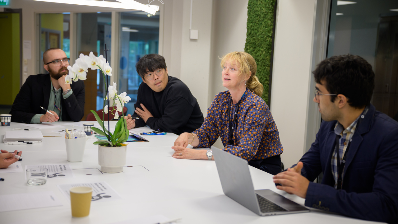 Four people sit at a table. The woman who is third from the left is speaking while the others, three men, listen.