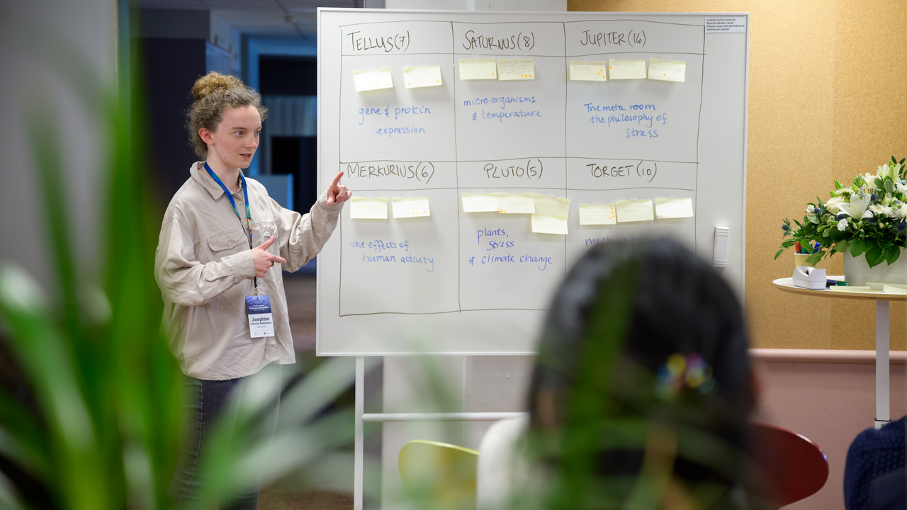 Josephine stands next to a whilte board with post-its and topics displayed in boxes.