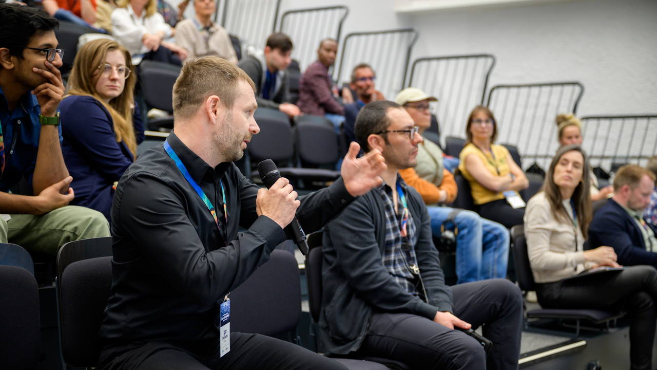 A man holds a microphone while seated in an auditorium with many other people behind him