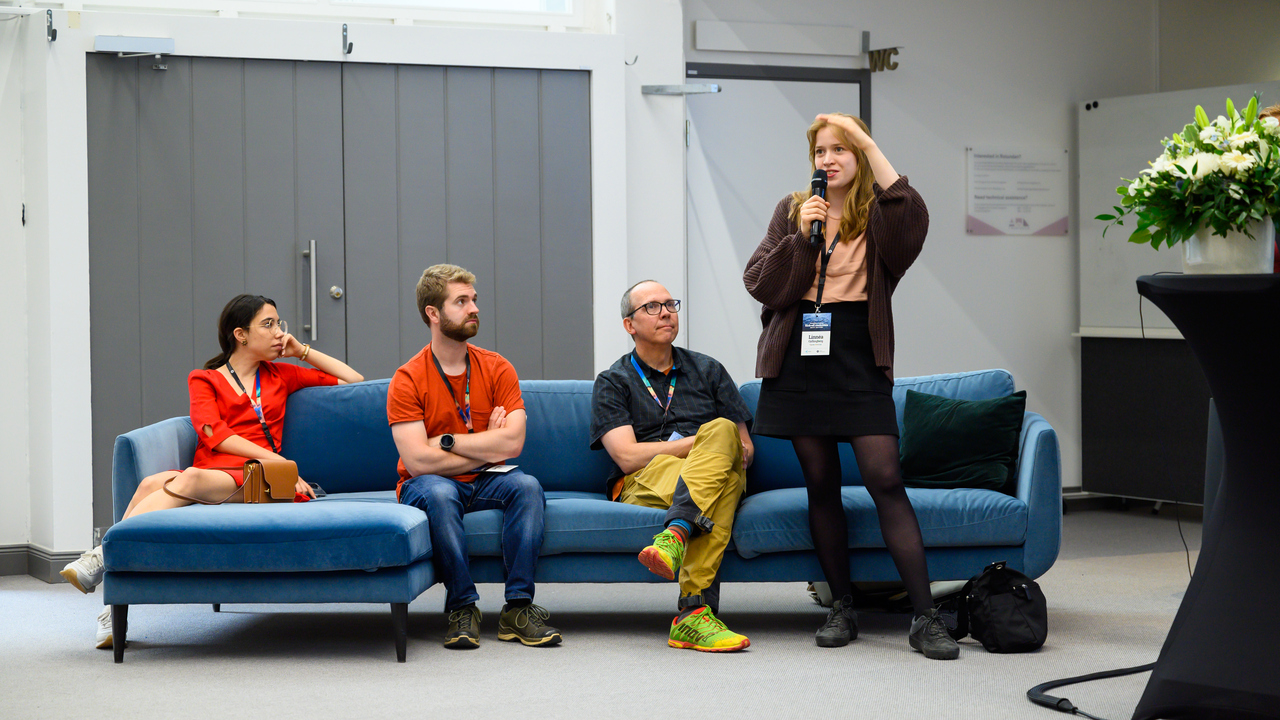 Tre personer sitter i en blå soffa och lyssnar på Linnea som står och talar medan hon håller i en mikrofon.