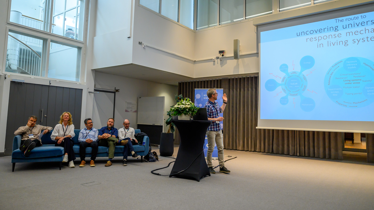 Five people sit on a blue sofa and one stands speaking next to a large screen.