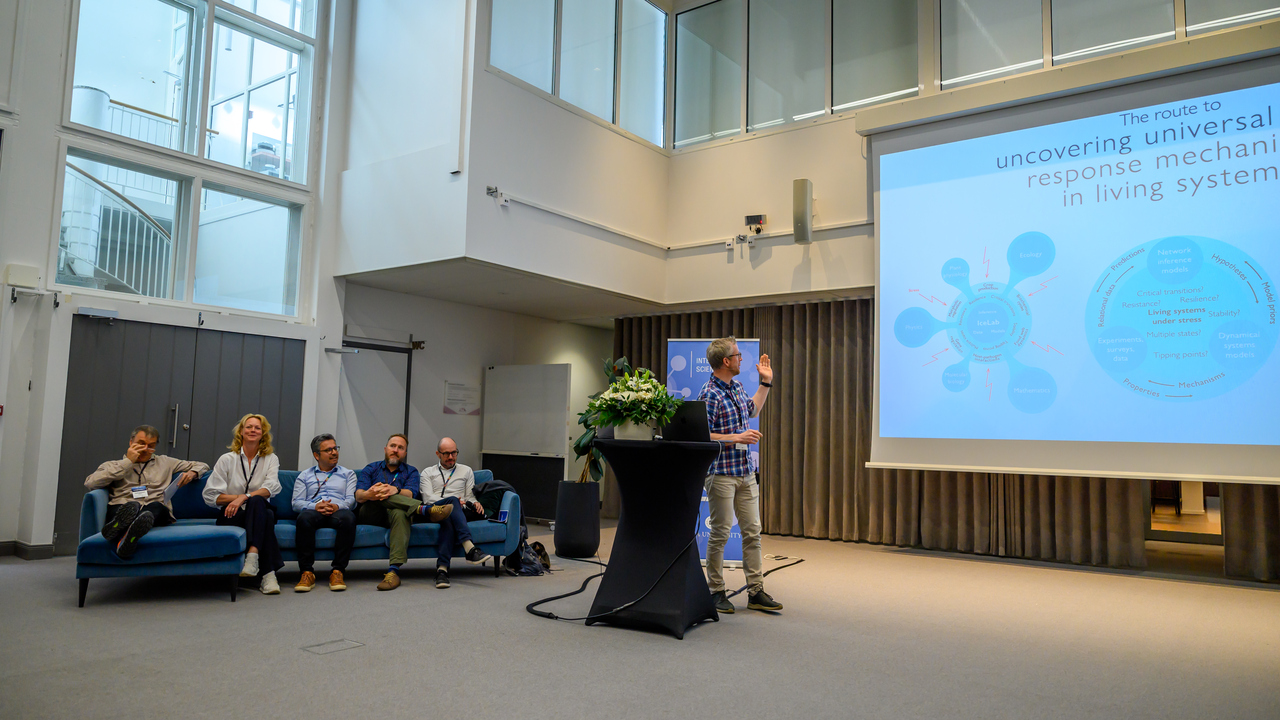 Fem personer sitter i en blå soffa och en står och talar bredvid en stor skärm.