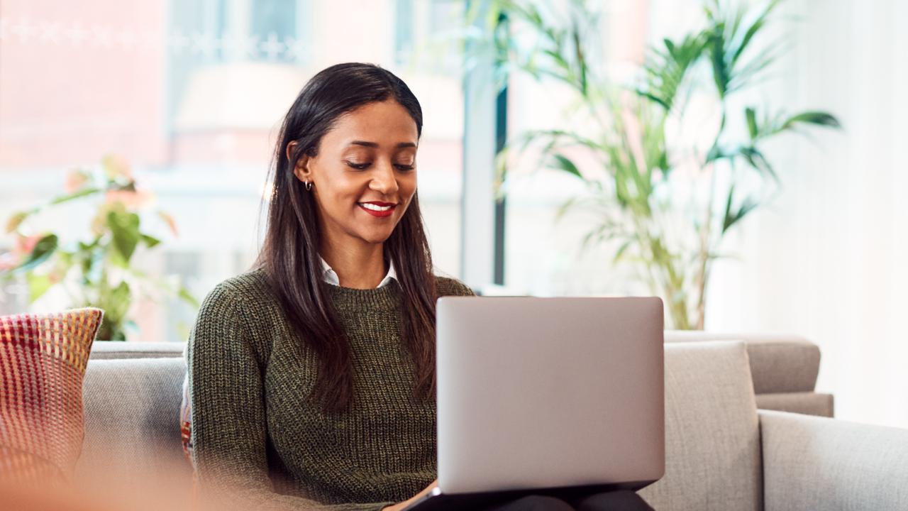 Glad kvinna sitter med laptop i beige soffa i kontorsmiljö.