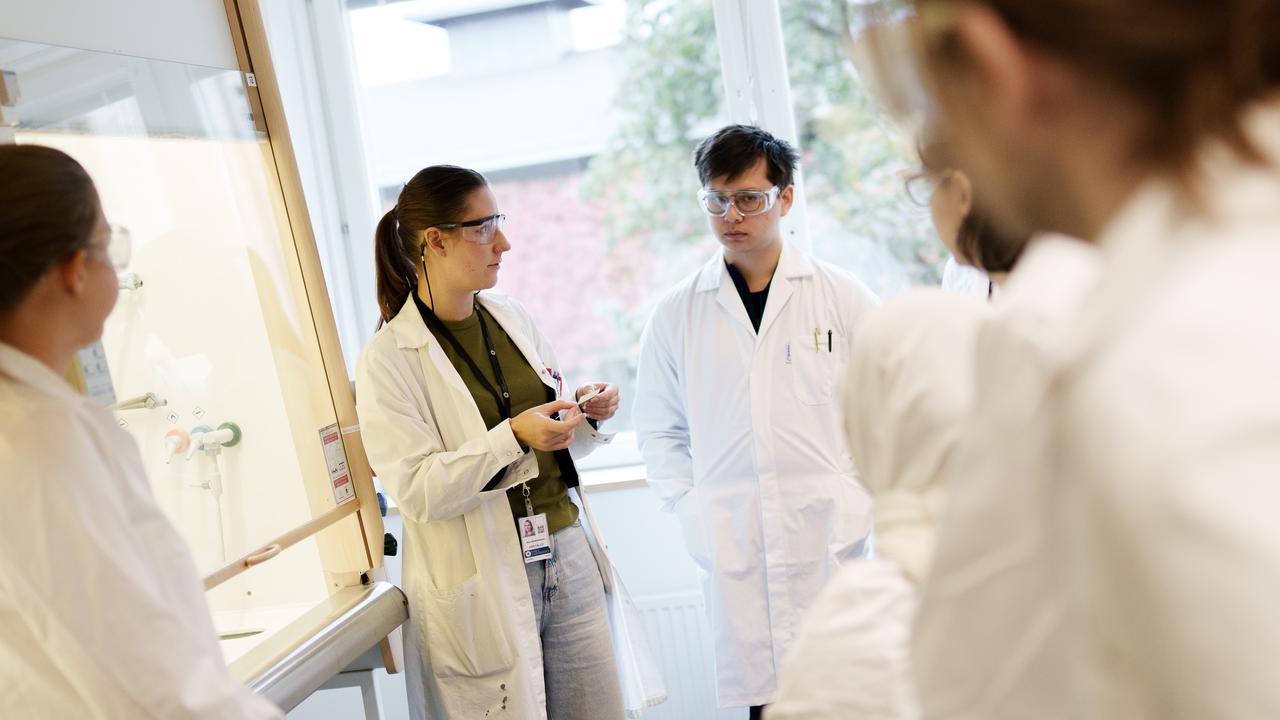 Bild på lärare och en grupp studenter i labbrockar under genomgång.