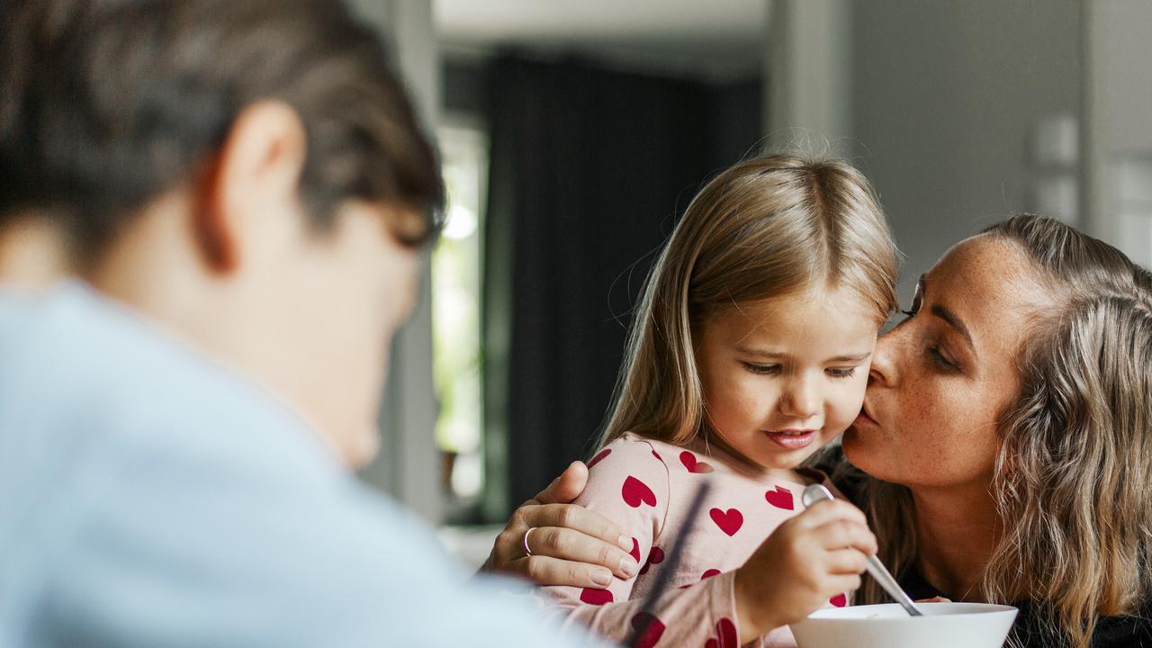 Mamma kysser sin dotter på kinden vid frukostbordet. En man syns i förgrunden.