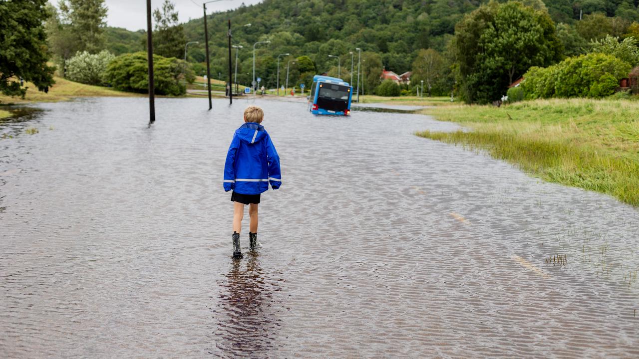 Ett barn står vänd mot en översvämmad bilväg. Längre fram syns en buss.