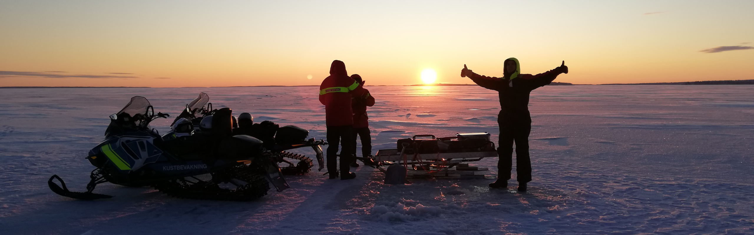 Tre personer står på havsis med skotrar och provtagningsutrustning medan solen knappt når över horisonten