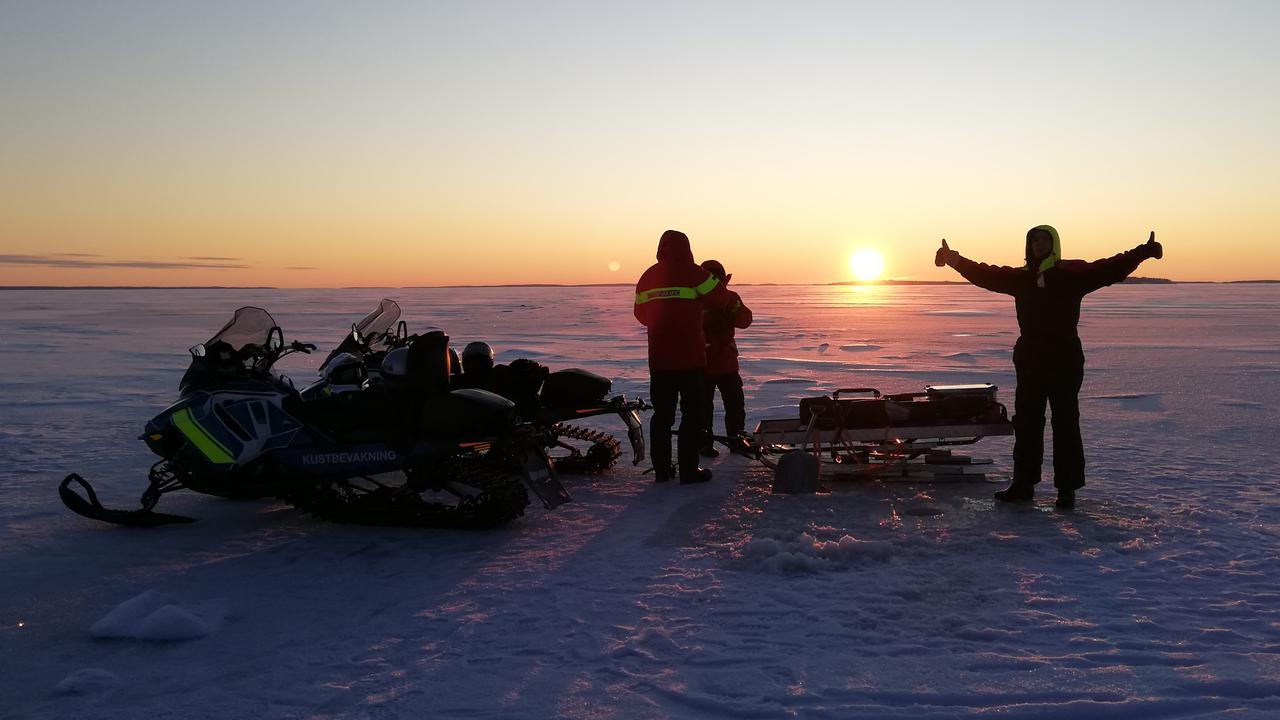 Tre personer står på havsis med skotrar och provtagningsutrustning medan solen knappt når över horisonten