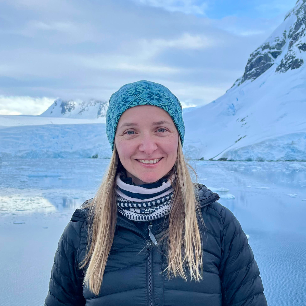 Kylie Owen står i mörkblå vinterjacka och ljusblå mössa och ler mot kameran. I bakgrunden syns en spegelblank sjö, snö och ett berg.