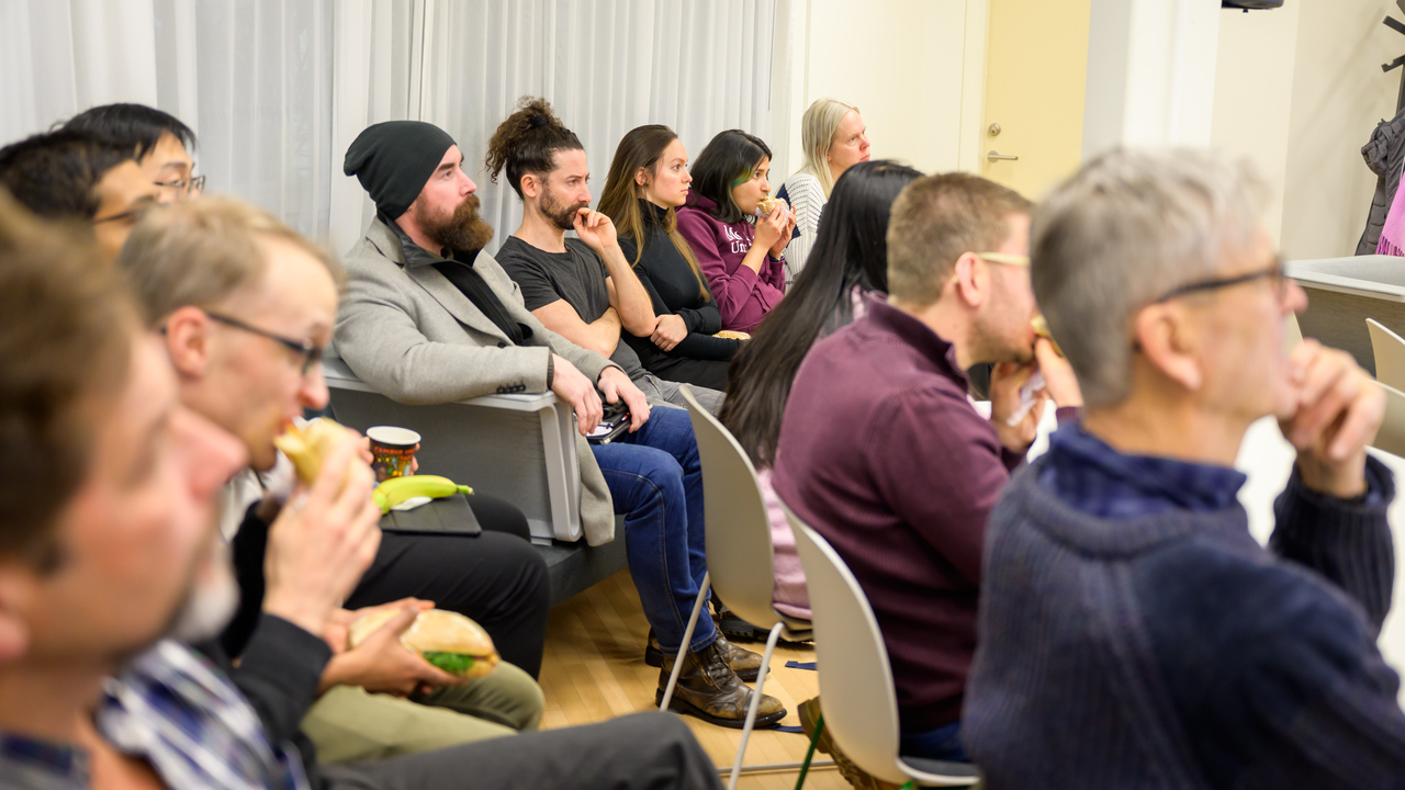 An audience of seated researchers listen intently to a research pitch while eating their lunches
