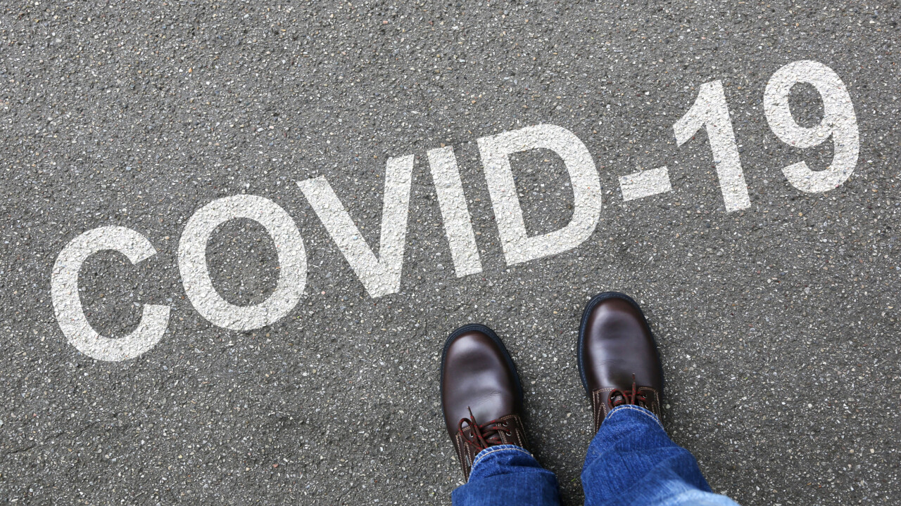 Fötter som står framför ordet Covid-19 skrivet på asfalten.