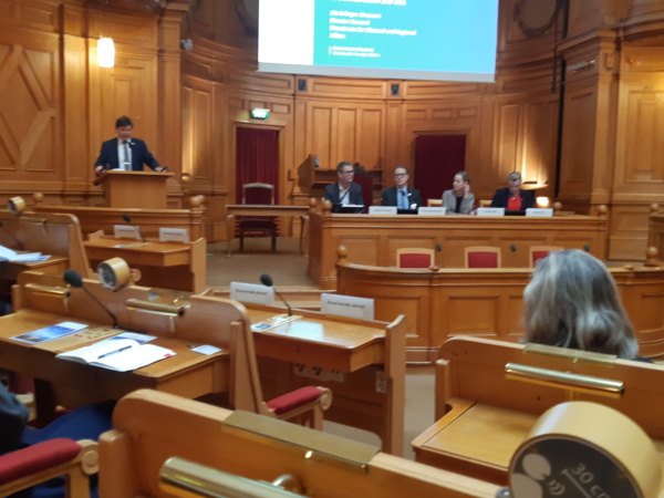 Människor sittandes vid ett presidium, och en ståendes vid podiet i Sveriges Riksdags andrakammare.