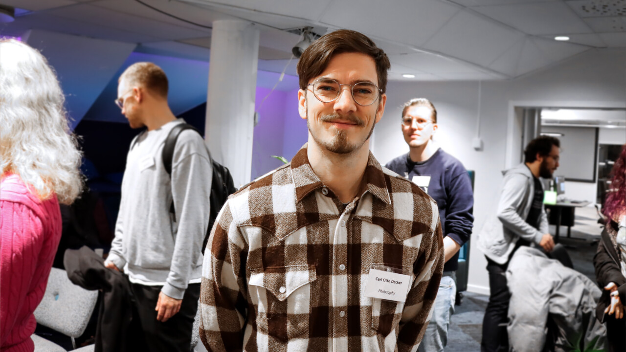 Bilden visar en manlig student, med mörkt hår och glasögon som tittar in i kameran. I bakgrunden syns andra studenter som deltog på Hackatonet.