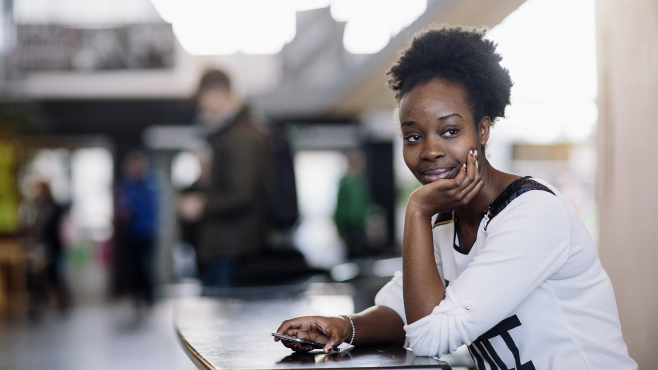 Kvinnlig student som sitter och funderar inomhus på campus med en mobiltelefon i handen
