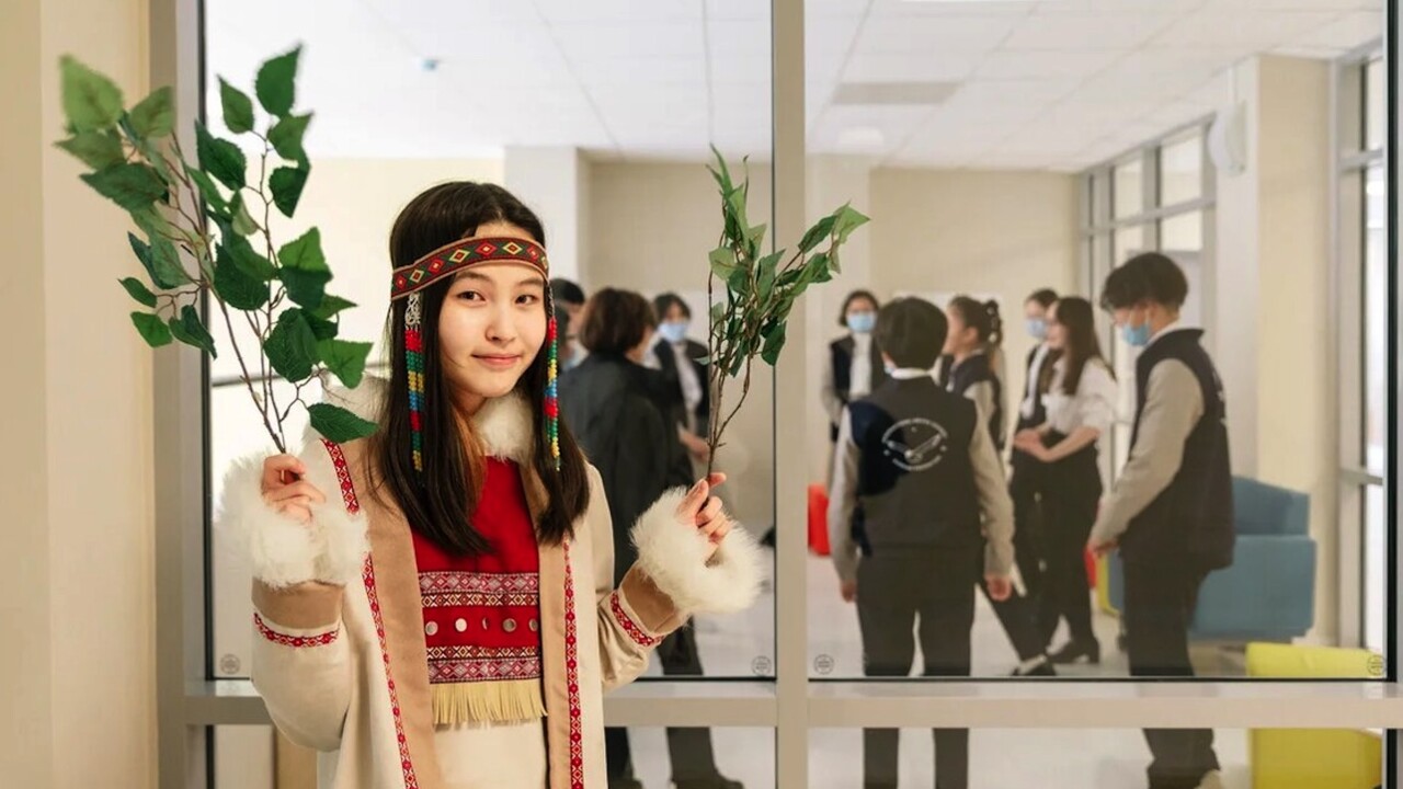 Student wearing traditional clothes holding up a leaf twig. Through the windows behind, more students and teachers can be seen gathering in a circle