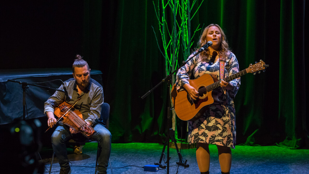 Bild på Carolina Miskovsky som sjunger och spelar gitarr och Daniel Pettersson som spelar nyckelharpa på scenen i Aula Nordica, Umeå universitet.
