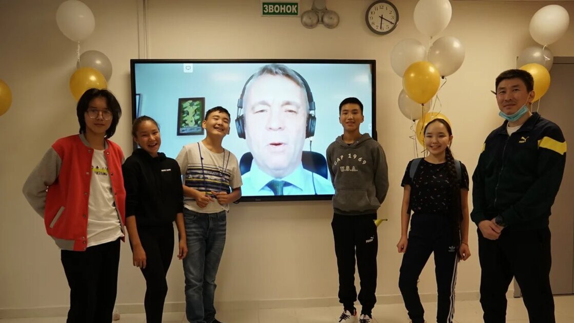 Elever står på vardera sida om en storbildsskärm om ler mot kameran medan Peter Skölds välönskningar syns på skärmen i bakgrunden