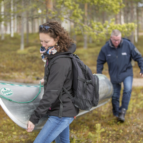 Två personer som bär en kanot genom skogen mot vattnet.