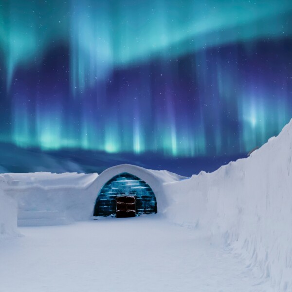A snow building with a wooden door, high snow edges and a dark blue sky with northern lights.