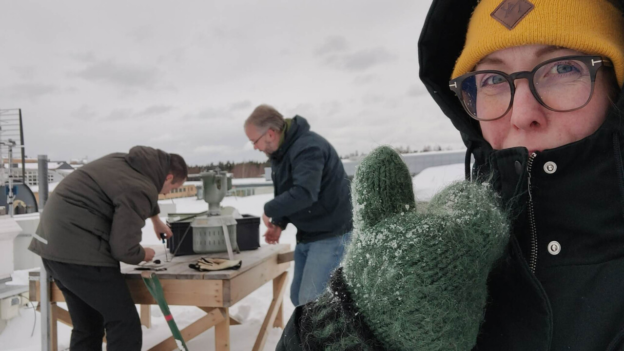 Två personer står vid ett bord på ett hustak böjda över en apparat i metall, en tredje person tittar in i kameran och gör tummen upp.