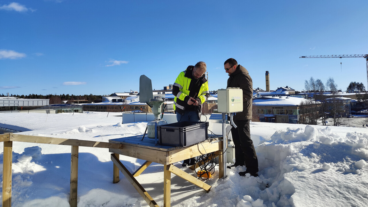 Två personer står vid ett bord på ett hustak böjda över en apparat i metall