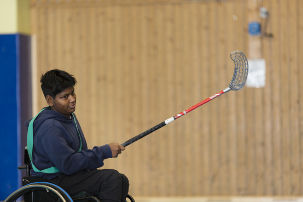 Kille i rullstol med innebandyklubba i handen skjuter iväg en boll.