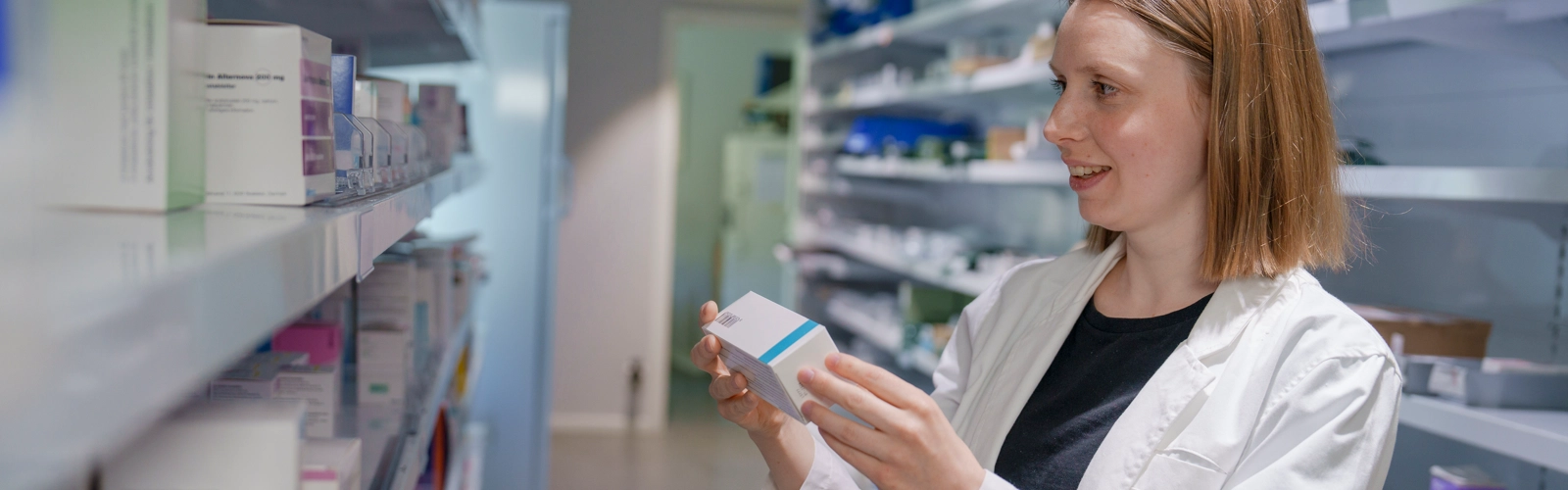 Apotekspersonal vid en hylla med läkemedelsförpackningar. Personen håller i en läkemedelsförpackning.