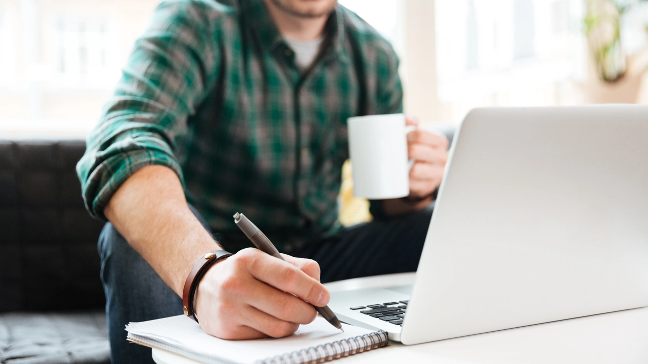 Person sitter i en soffa och arbetar. Har dator, kaffekopp och block där han skriver.