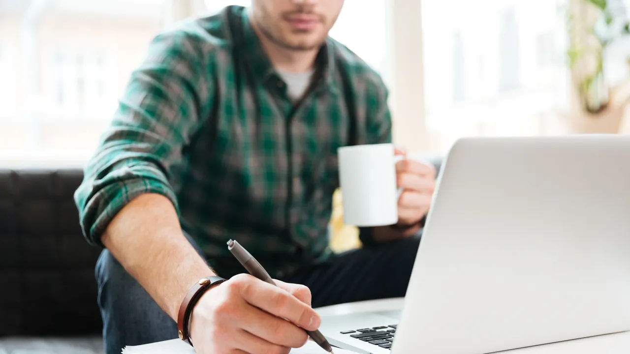 Man som sitter framför en dator med en kaffekopp och skriver med en penna.