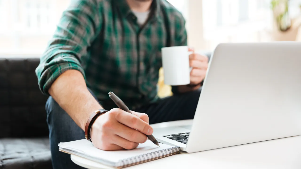 Person sitter i en soffa och arbetar. Har dator, kaffekopp och block där han skriver.