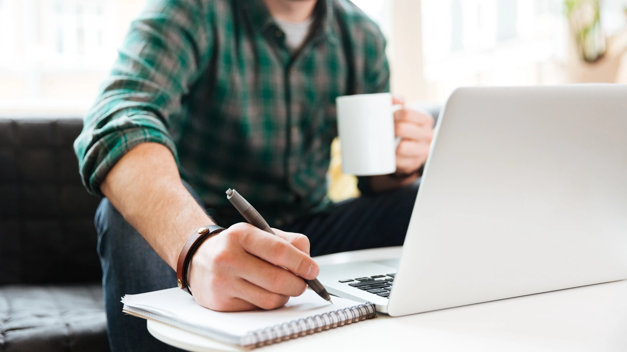 Person sitting on a sofa working. Laptop, coffee and notepad in front of him.