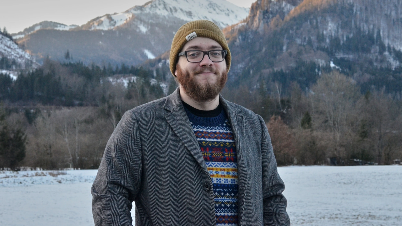 Photo of Bastian Schiffthaler standing outdoors, in the background lake, forest and snowcovered mountain tops