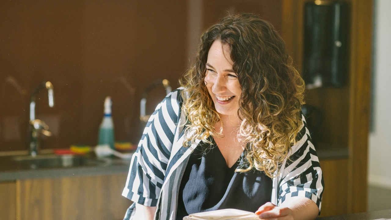 En leende kvinna äter sin lunchlåda i ett gemensamt lunchrum.