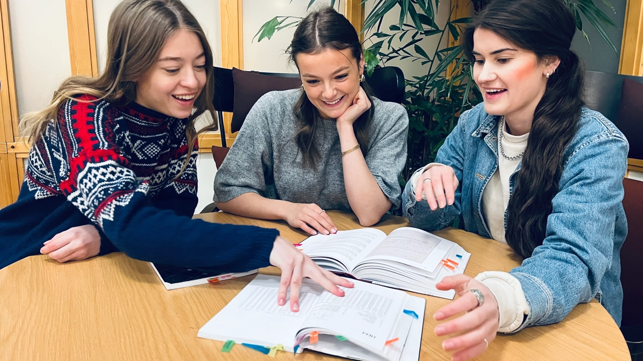 Bild på studenterna Eira, Olivia och Elsa vid ett bord med kursböcker som de bläddrar i