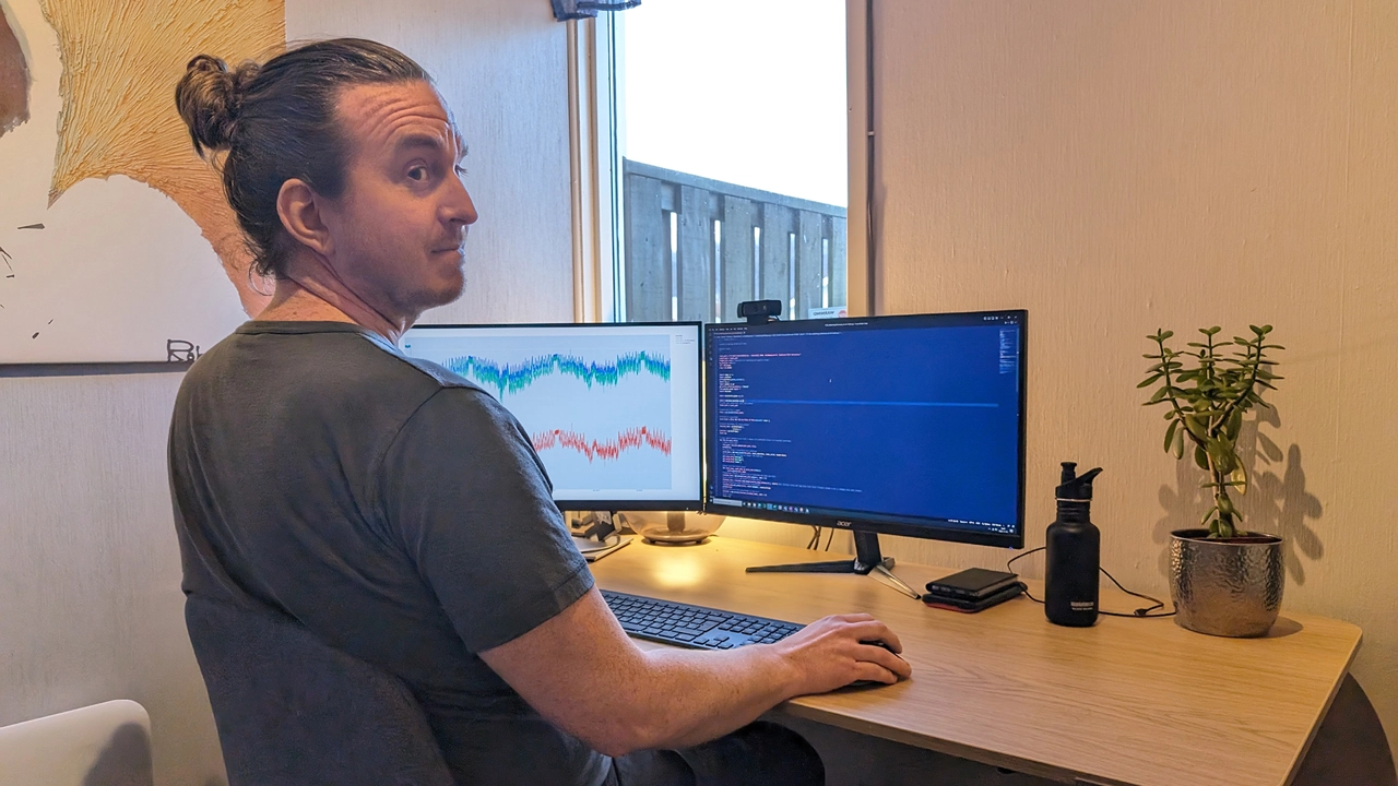 Bild på Daniel Dahlstrand sedd snett bakifrån, stående vid skrivbord med två datorskärmar