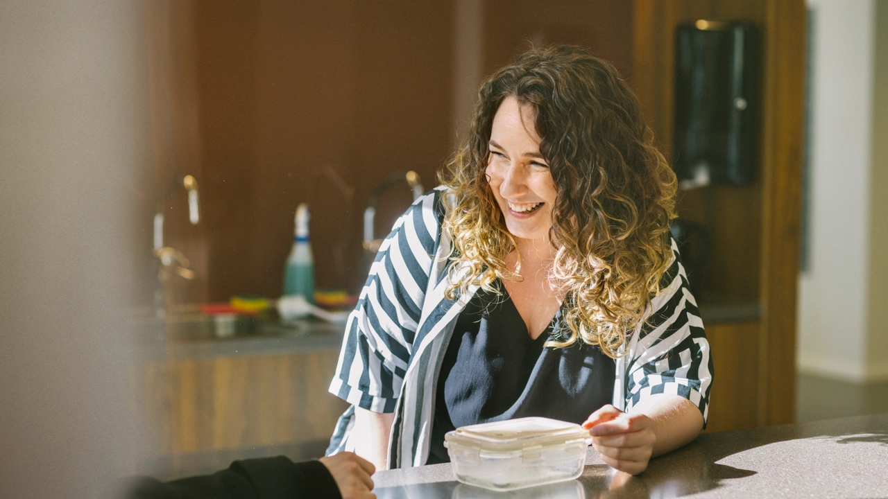 En leende kvinna äter sin lunchlåda i ett gemensamt lunchrum.