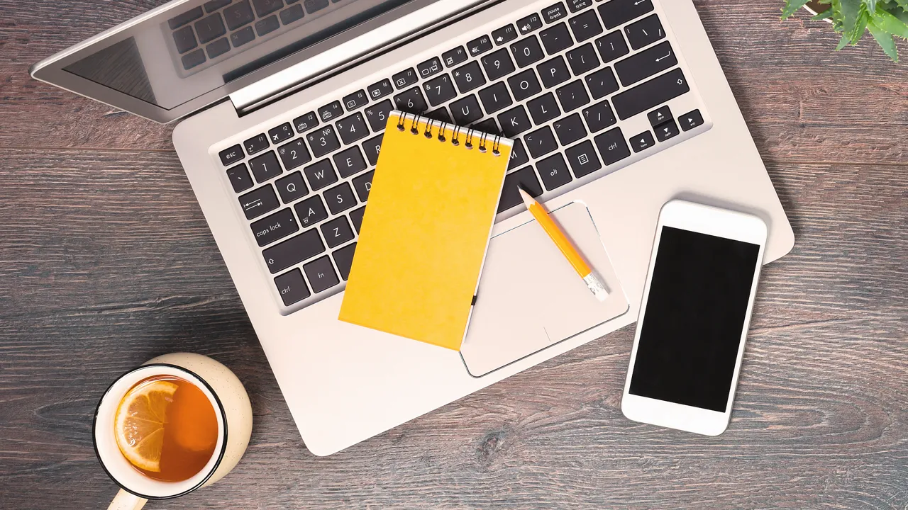 Dator, anteckningsblock, mobil och penna fotat uppifrån på ett bord.