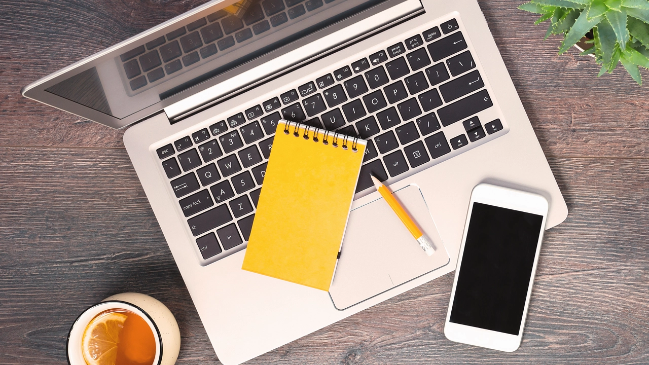 Dator, anteckningsblock, mobil och penna fotat uppifrån på ett bord.