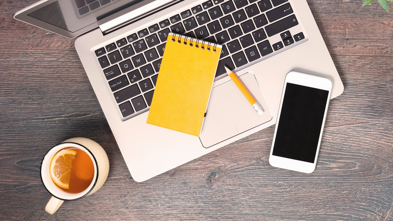 Dator, anteckningsblock, mobil och penna fotat uppifrån på ett bord.