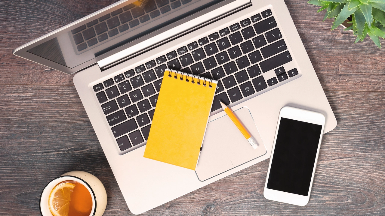 Dator, anteckningsblock, mobil och penna fotat uppifrån på ett bord.